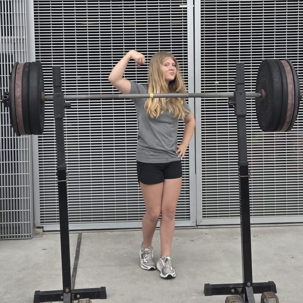 Maddy (Pre-Teen Girl) Showing Off Her Bicep with A Barbell in Front of Her